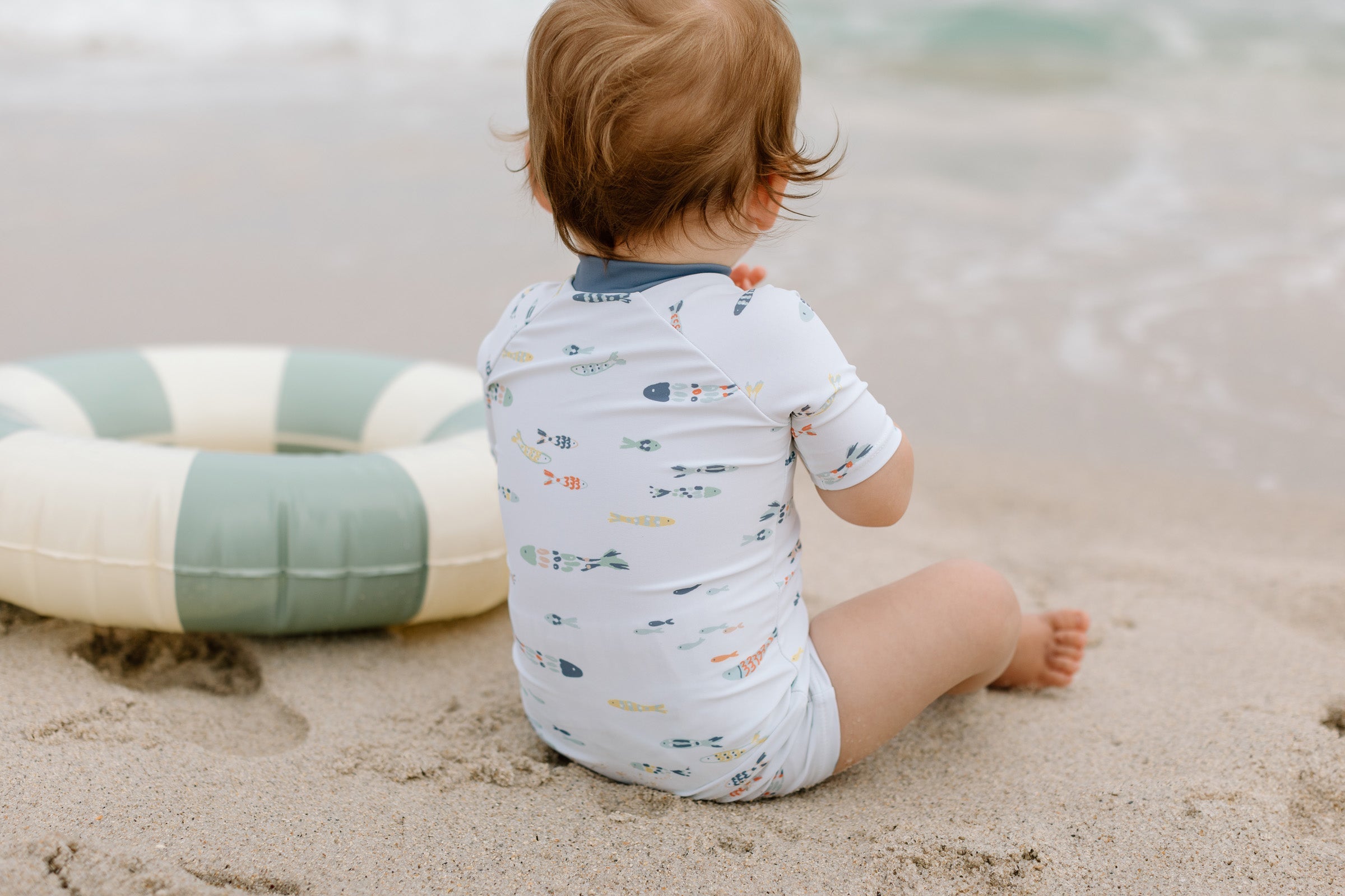 A baby sit on the sand in a Pehr swimsuit.